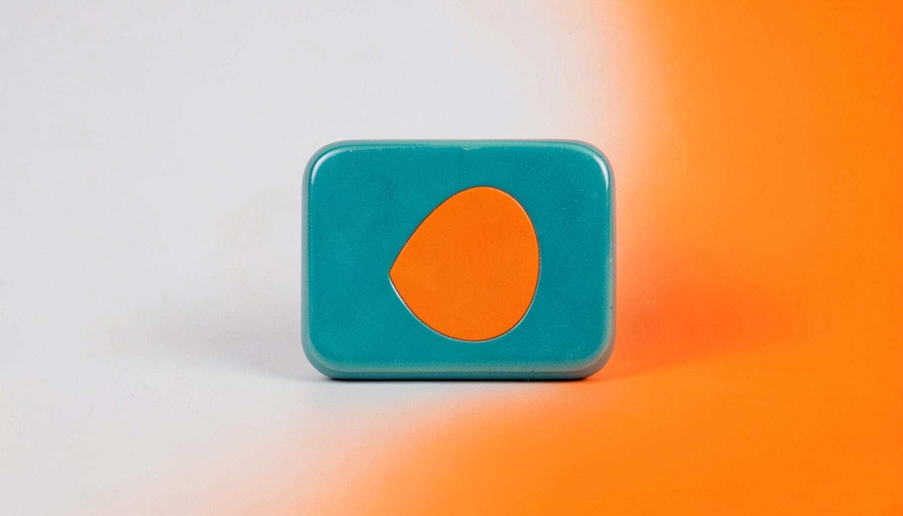 A crisp studio photograph on a seamless light grey backdrop of a rounded tile in teal with an abstract orange accent shape, evenly lit.